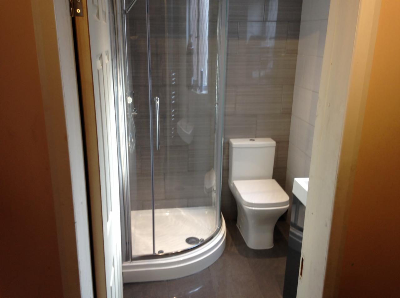 Wykończona łazienka z kabiną prysznicową narożną, toaletą kompaktową i szarą glazurą imitującą drewno, widok zza otwartych drzwi.