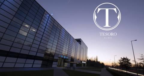 Nowoczesny budynek kliniki z szklaną fasadą oświetlony zachodzącym słońcem, z widocznym logo 'Tesoro Clinic' w prawym górnym rogu i alejką prowadzącą do wejścia.