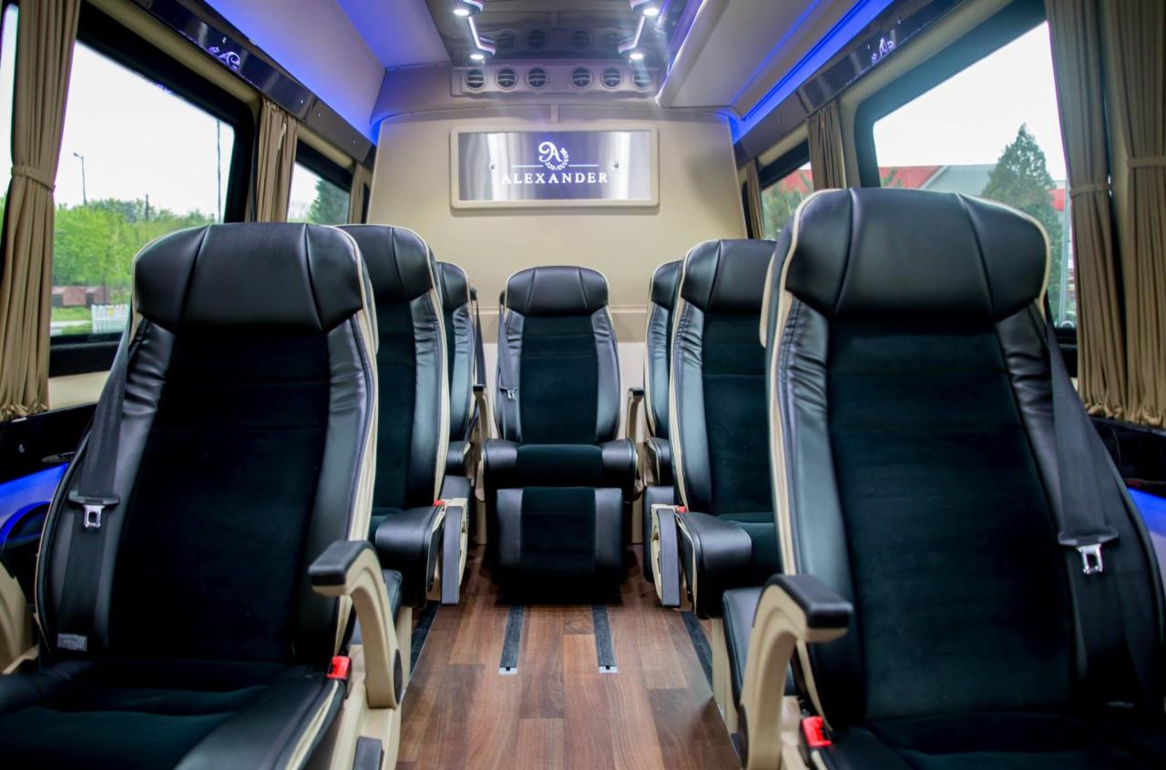 Wnętrze luksusowego busa z czarnymi skórzanymi fotelami, drewnianą podłogą i subtelnym niebieskim oświetleniem LED pod sufitem, widoczne okna z zasłonami i tabliczka z nazwą 'ALEXANDER'...