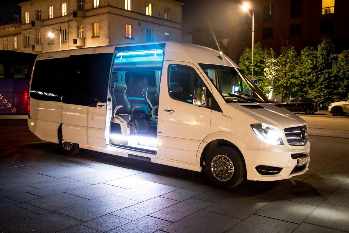 Biały, luksusowy bus Mercedes-Benz VIP Line z otwartymi drzwiami przesuwnymi, oświetlony od wewnątrz, zaparkowany nocą na brukowanej ulicy z budynkami w tle.