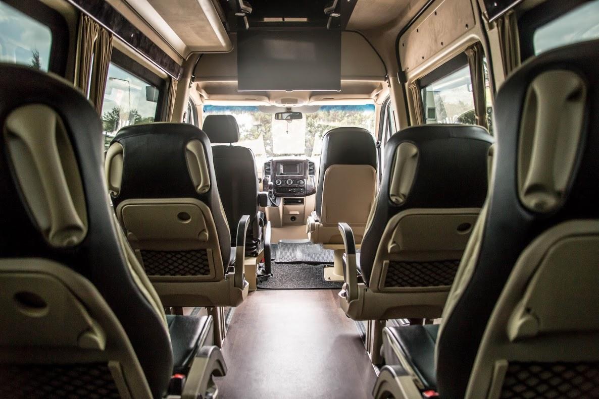 Wnętrze komfortowego busa pasażerskiego z czarnymi skórzanymi fotelami, siatkowymi kieszeniami i zamontowanym ekranem, widok z tyłu w kierunku kabiny kierowcy.