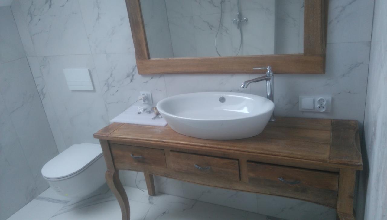 Biała umywalka nablatowa na drewnianej szafce z szufladami w łazience wykończonej marmurowymi płytkami. W tle lustro w drewnianej ramie i toaleta.