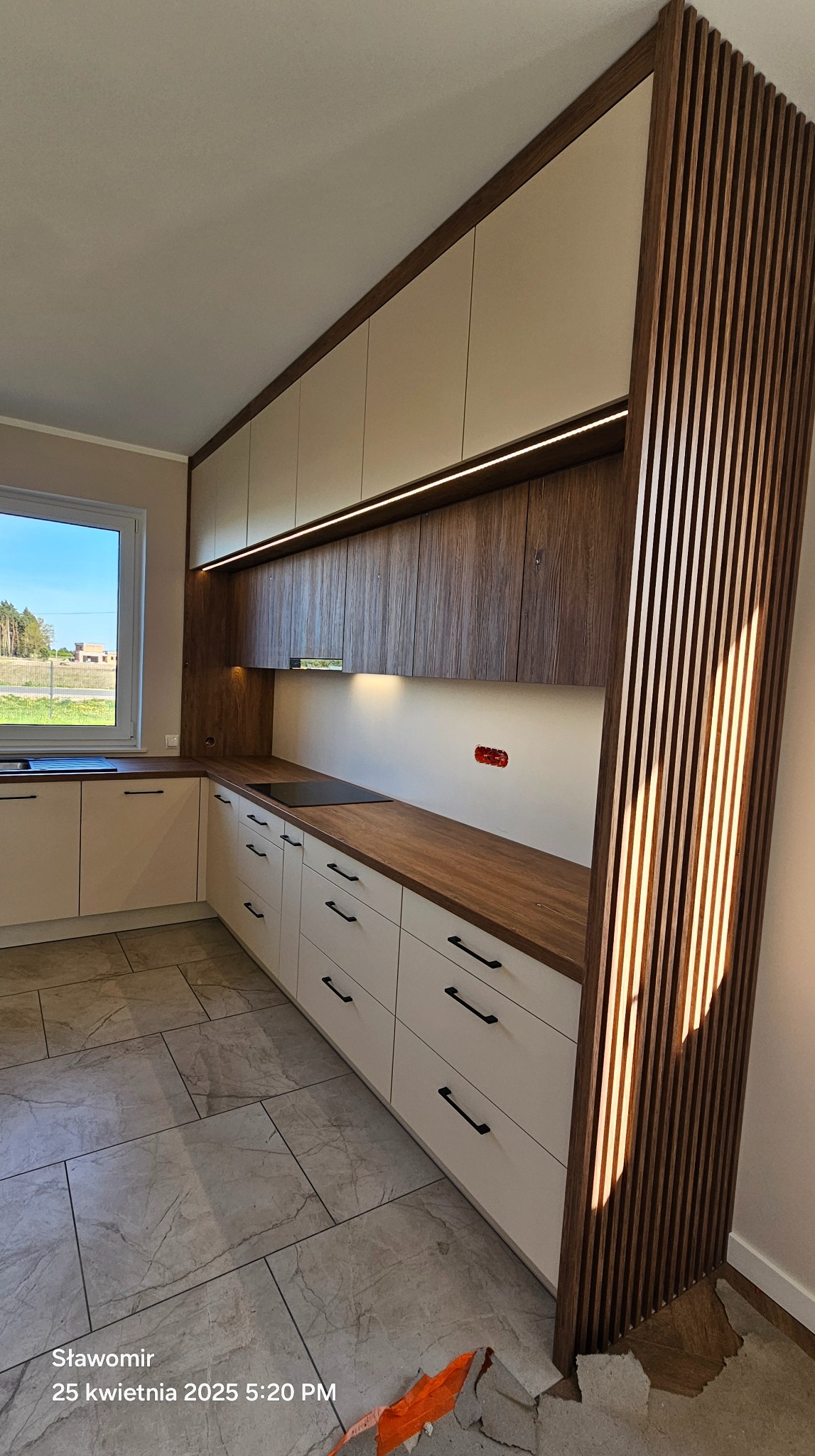 Nowoczesna kuchnia z drewnianymi akcentami: blaty i pionowe lamele. Białe fronty szafek, czarne uchwyty, oświetlenie LED pod szafkami wiszącymi. Widok na okno.