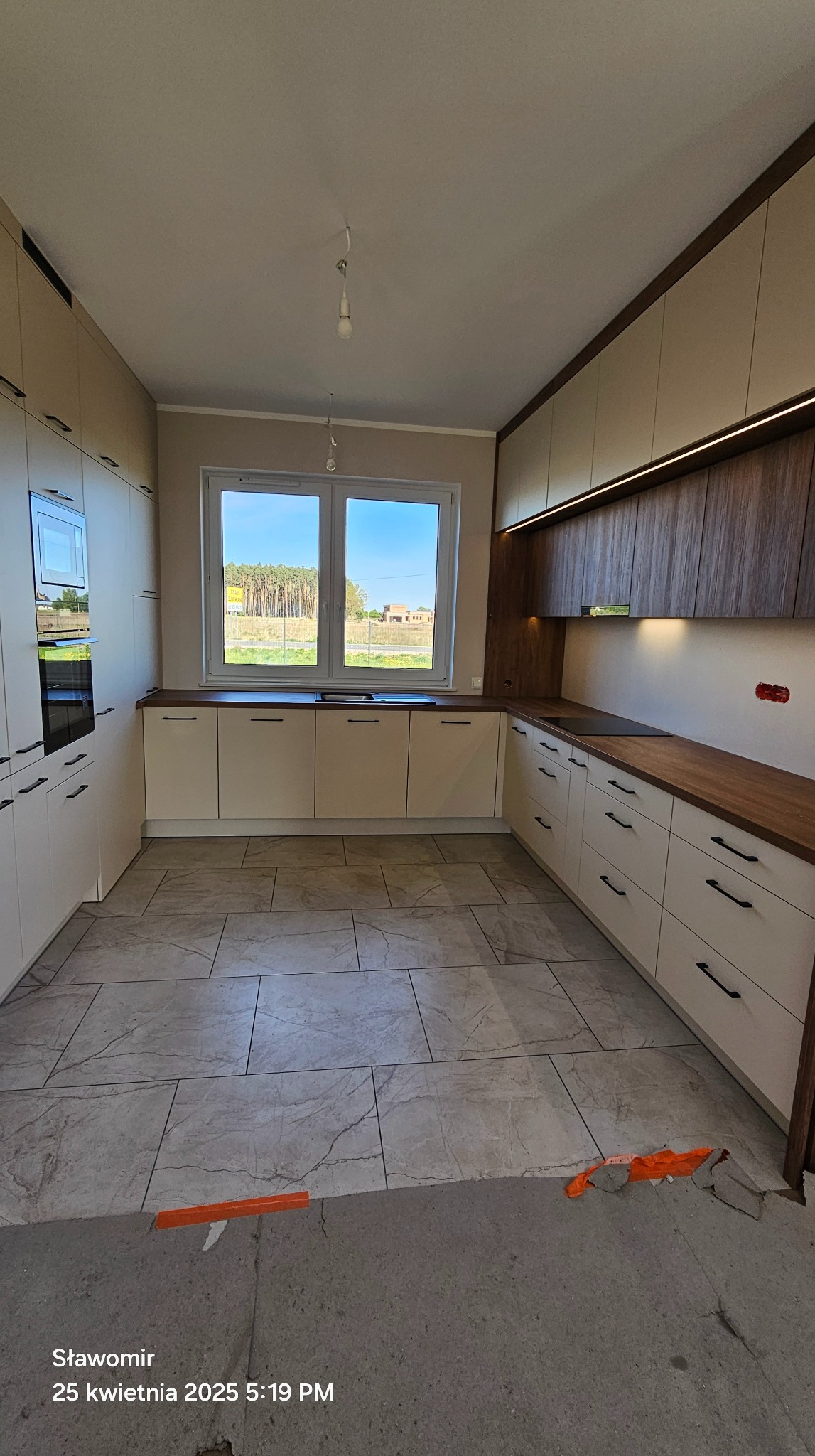 Jasna kuchnia z drewnianym blatem i oświetleniem LED podszafkowym. Widok na okno z zielenią. Minimalistyczne uchwyty. Podłoga wyłożona płytkami imitującymi marmur.