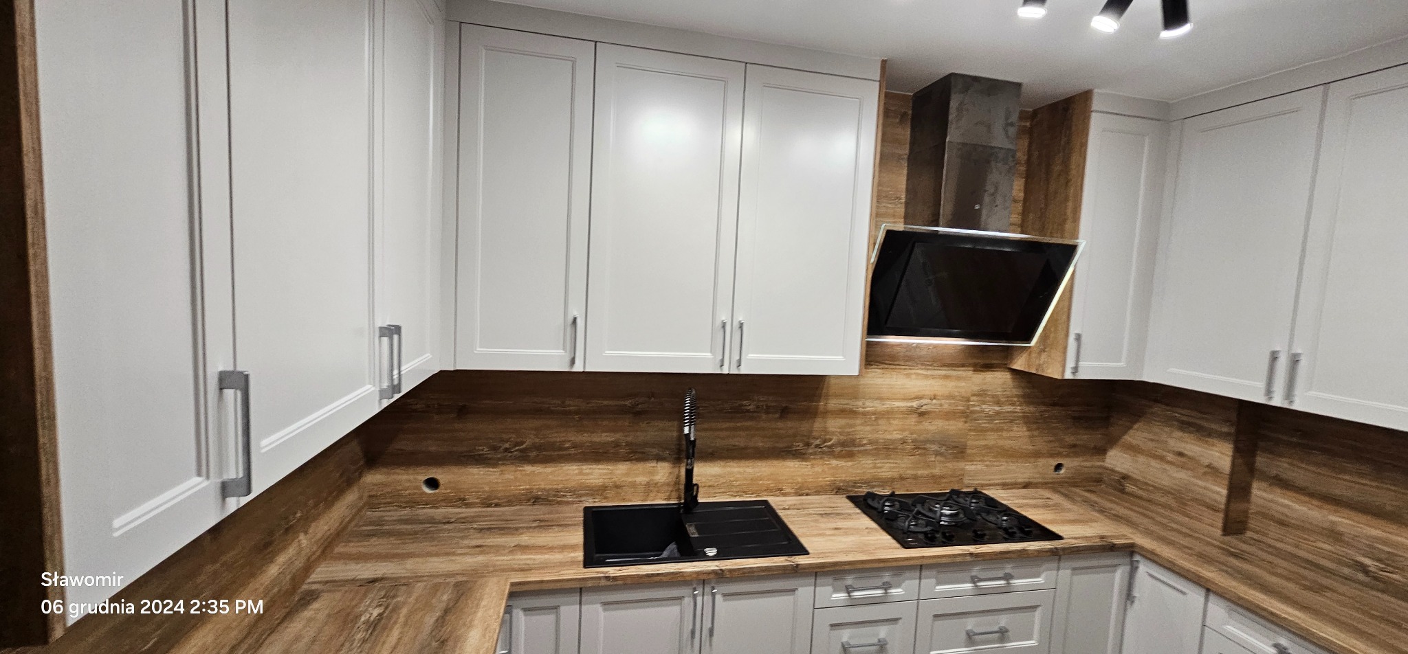 Nowoczesna kuchnia z białymi szafkami i drewnianym blatem. Czarny zlew i płyta grzewcza. Uwagę przyciąga drewniana ściana nad blatem, dopasowana do blatu.