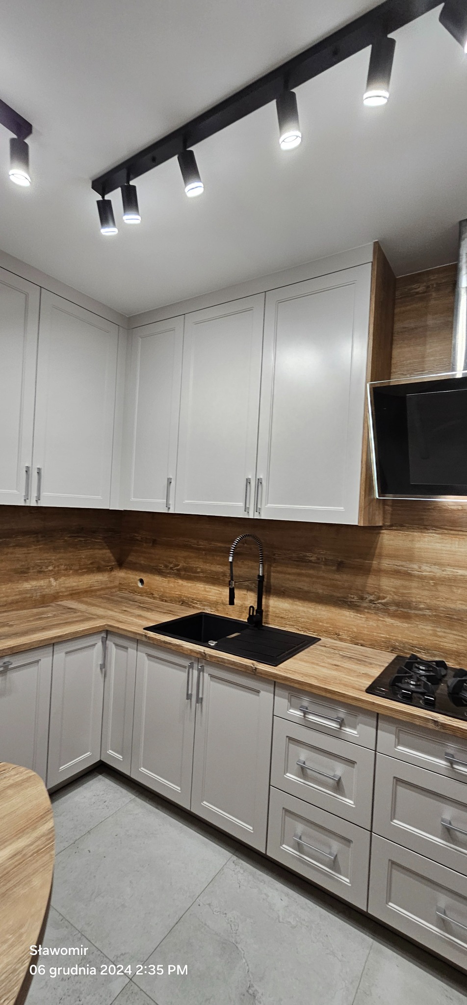 Jasna kuchnia z drewnianym blatem i czarnym zlewem. Górne szafki białe, dolne szare. Oświetlenie szynowe. Nowoczesny design.