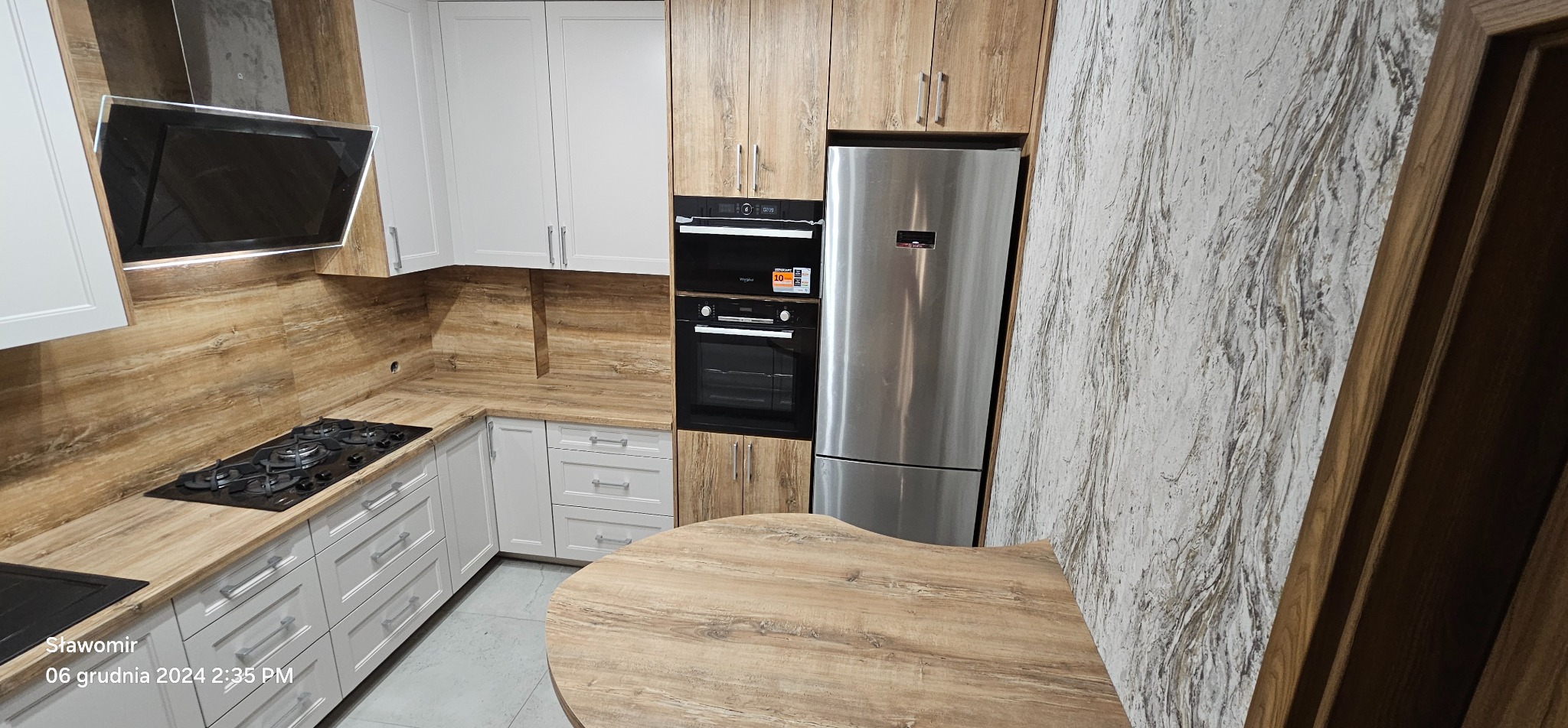 Jasna kuchnia z drewnianymi akcentami: białe szafki, drewnopodobne blaty i okładziny, czarna płyta grzewcza i piekarnik, srebrna lodówka. Nowoczesny design.