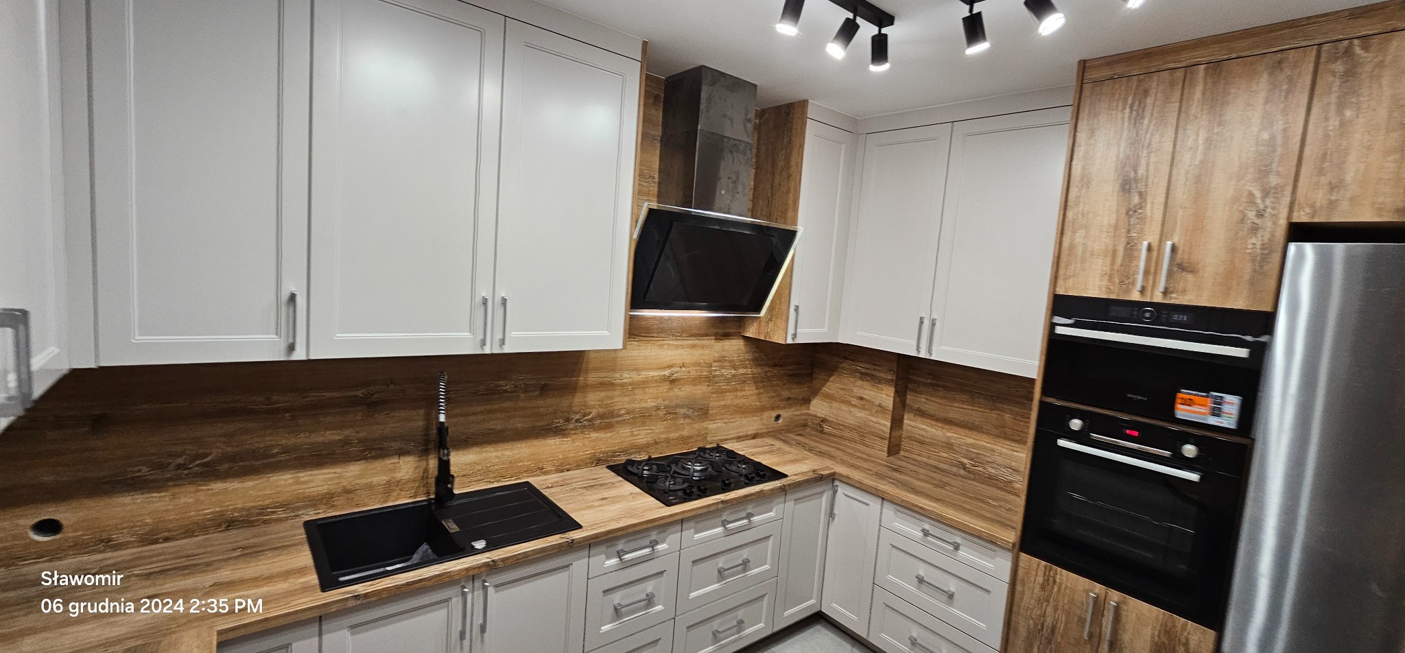 Nowoczesna kuchnia na wymiar z białymi szafkami i drewnianym blatem. Czarny zlew i płyta grzewcza. Oświetlenie punktowe. Realizacja z dbałością o detale.