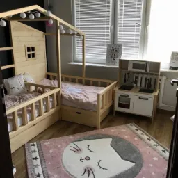 Pokój dziecięcy z drewnianym łóżkiem w kształcie domku, oświetlonym girlandą z kul, obok drewniana kuchnia dla dzieci, na podłodze różowy dywan z motywem kota.