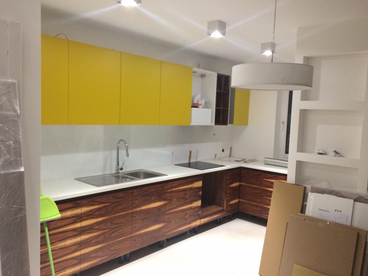 Nowoczesna kuchnia z żółtymi szafkami górnymi i dolnymi szafkami z egzotycznego drewna o wyraźnym usłojeniu, biały blat i ściana nad blatem, widoczne elementy montażowe i kartony po sprzęcie.