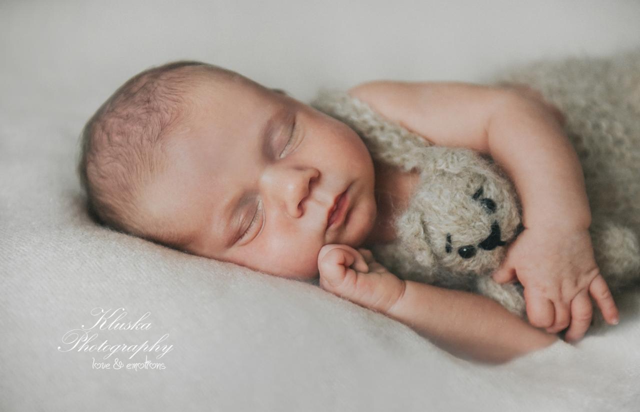 Śpiący noworodek w beżowym ubranku, przytulający pluszowego misia na kremowym tle, delikatne oświetlenie.