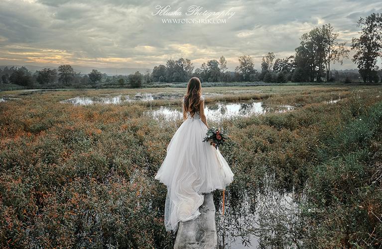 Panna młoda w białej sukni, z bukietem w ręku, stoi tyłem na drewnianej kładce nad wodą, otoczona zielenią i trzcinami, w Dąbrowie Górniczej, w tle widoczne drzewa i pochmurne niebo.