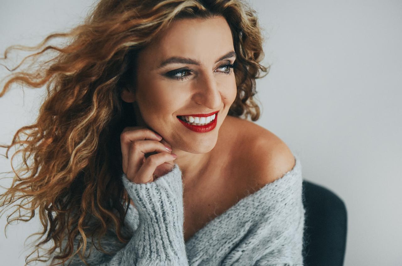 Uśmiechnięta kobieta z burzą blond loków, w szarym swetrze z odkrytym ramieniem, z ręką przy twarzy, spogląda w bok.