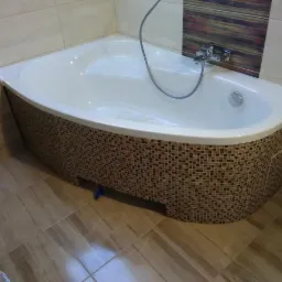 Biała wanna narożna z mozaikową obudową w odcieniach brązu, z chromowaną baterią i słuchawką prysznicową, na tle beżowych płytek ściennych i podłogowych, z widocznym fragmentem toalety.