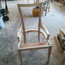 Niedokończony drewniany szkielet krzesła w warsztacie stolarskim, widoczne połączenia elementów i surowe drewno, zarys innych mebli w tle.