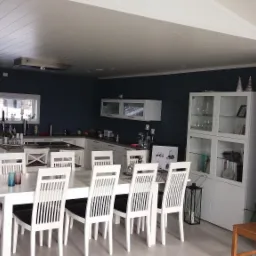 Jasna kuchnia z ciemnymi ścianami, białe meble i stół z krzesłami, sufit wykończony panelami, widoczny fragment antresoli.