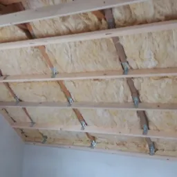 Izolacja z wełny mineralnej zamontowana między drewnianymi belkami stropu poddasza, widoczne metalowe łączniki, fragment ściany w tle.