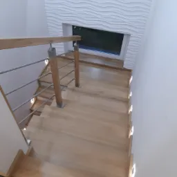 Drewniane schody z metalową balustradą i oświetleniem LED w stopniach, widok z góry, obok ściana z dekoracyjną fakturą i okno.