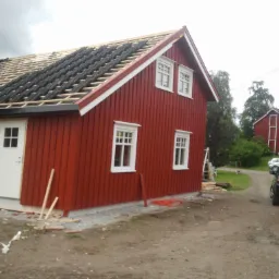 Czerwony dom z drewnianą elewacją w trakcie wymiany dachu, widoczne krokwie i częściowo ułożona dachówka, na podwórku zaparkowana przyczepa ze sprzętem.