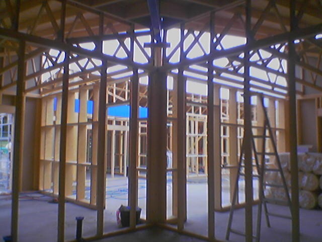 Wnętrze domu w trakcie budowy, widoczna drewniana konstrukcja szkieletowa ścian działowych i stropu, drabina oparta o ścianę, oraz rolki materiału izolacyjnego w tle.