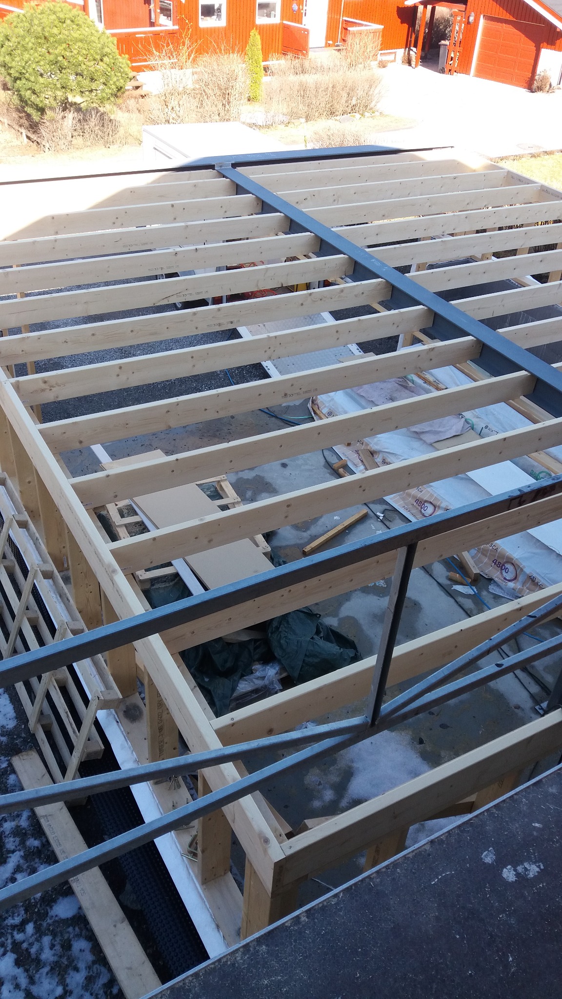 Konstrukcja drewniana z elementami stalowymi, prawdopodobnie w trakcie budowy domu, widok z góry na szkielet dachu i ścian, w tle czerwony budynek.