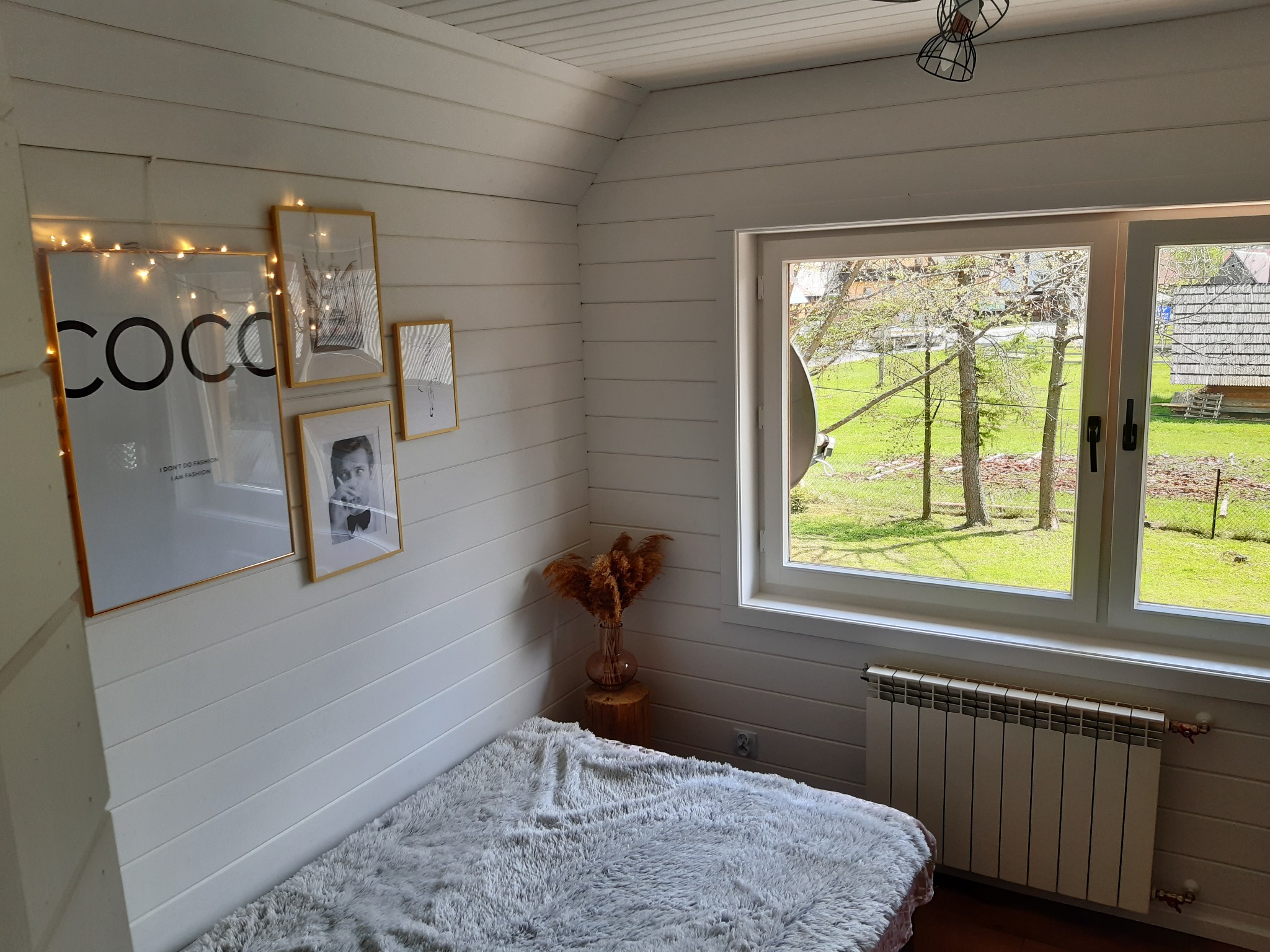 Poddasze wykończone białą boazerią z dekoracjami: plakat Coco Chanel z lampkami, inne grafiki w złotych ramkach, pampasy w wazonie na pniu, futrzany materac, okno z widokiem na wiejski krajobraz...