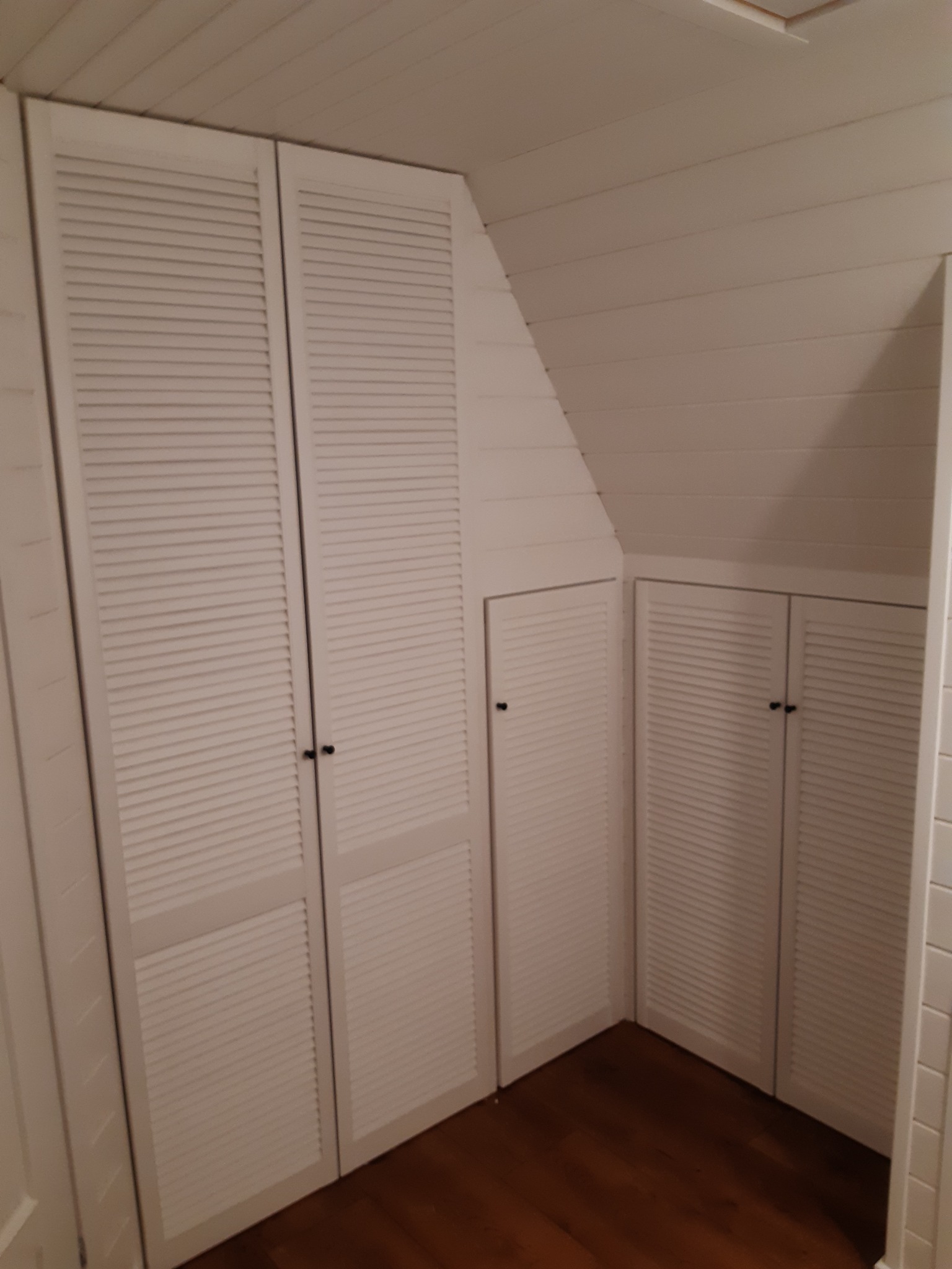 Białe, żaluzjowe drzwi szafy wnękowej pod skosem, z czarnymi uchwytami, w pokoju z drewnianą podłogą i boazerią.