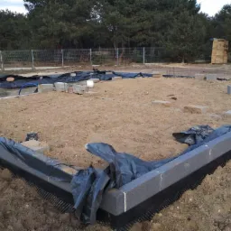 Przygotowanie fundamentów pod budowę domu: izolacja przeciwwilgociowa, bloczki betonowe i piasek na działce budowlanej.