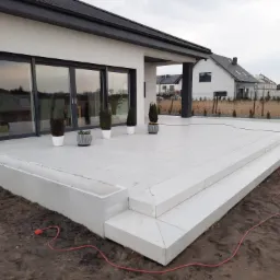 Nowoczesny taras z jasnych płyt z betonu architektonicznego, schody dwustopniowe, donice z iglakami, widok na dom z dużymi oknami przesuwnymi i ogród w tle, kabel elektryczny leżący na ziemi.