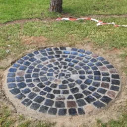 Okrągła nawierzchnia z ciemnej kostki ułożonej spiralnie, otoczona piaskiem i trawnikiem, widoczna taśma ostrzegawcza w tle, drzewo obok.