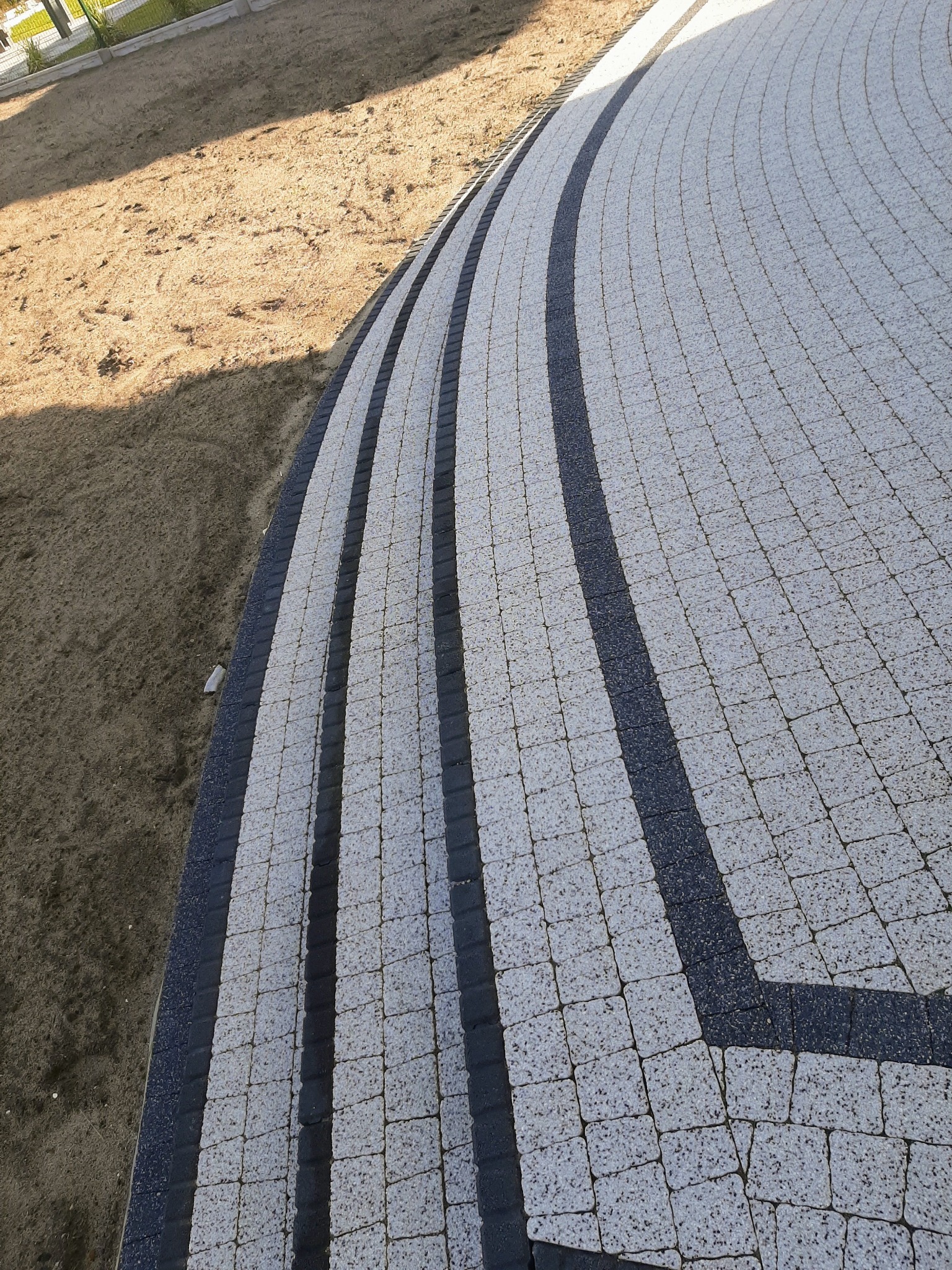 Zakrzywiona ścieżka z jasnoszarej kostki z ciemnymi pasami oddzielającymi, ułożona na przygotowanym podłożu z piasku.