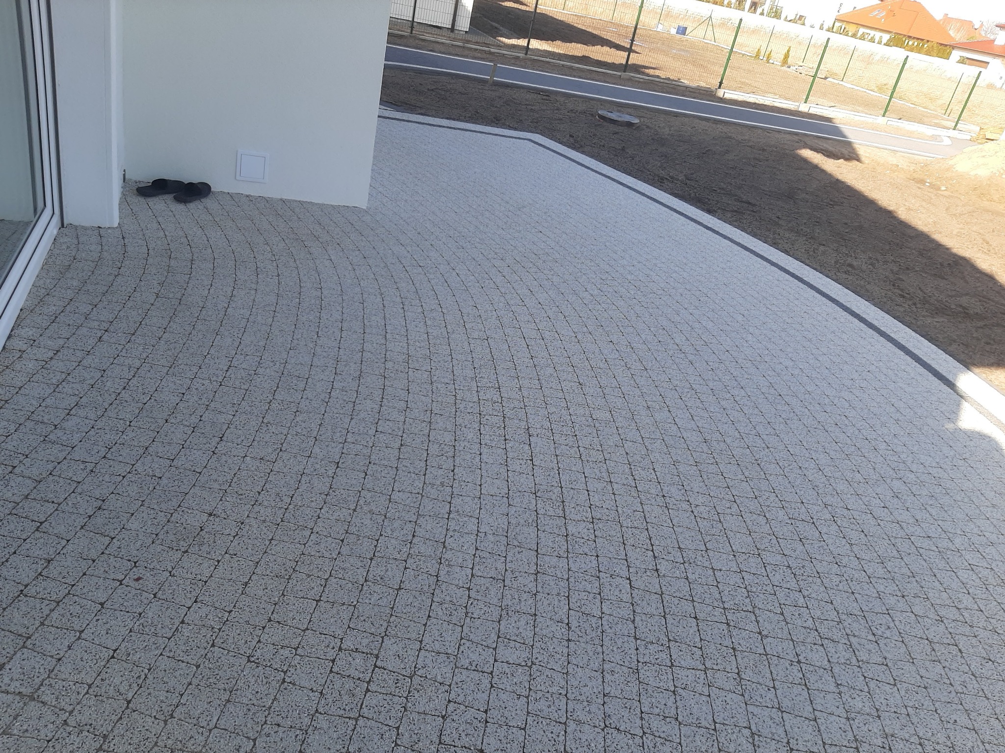 Jasnoszara kostka granitowa ułożona w łukowaty wzór na patio w Siekierkach Wielkich, z parą ciemnych klapek przy białej ścianie budynku.