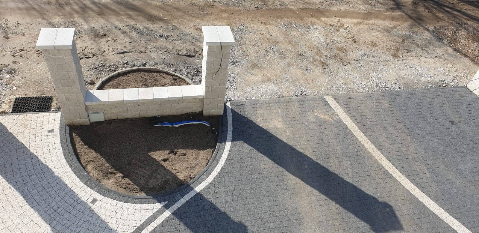Wysoka perspektywa na nowo wybrukowany podjazd z kostki brukowej w odcieniach szarości i bieli, z elementami dekoracyjnymi w postaci dwóch kamiennych słupków i zaokrąglonego rabatu z ziemią...