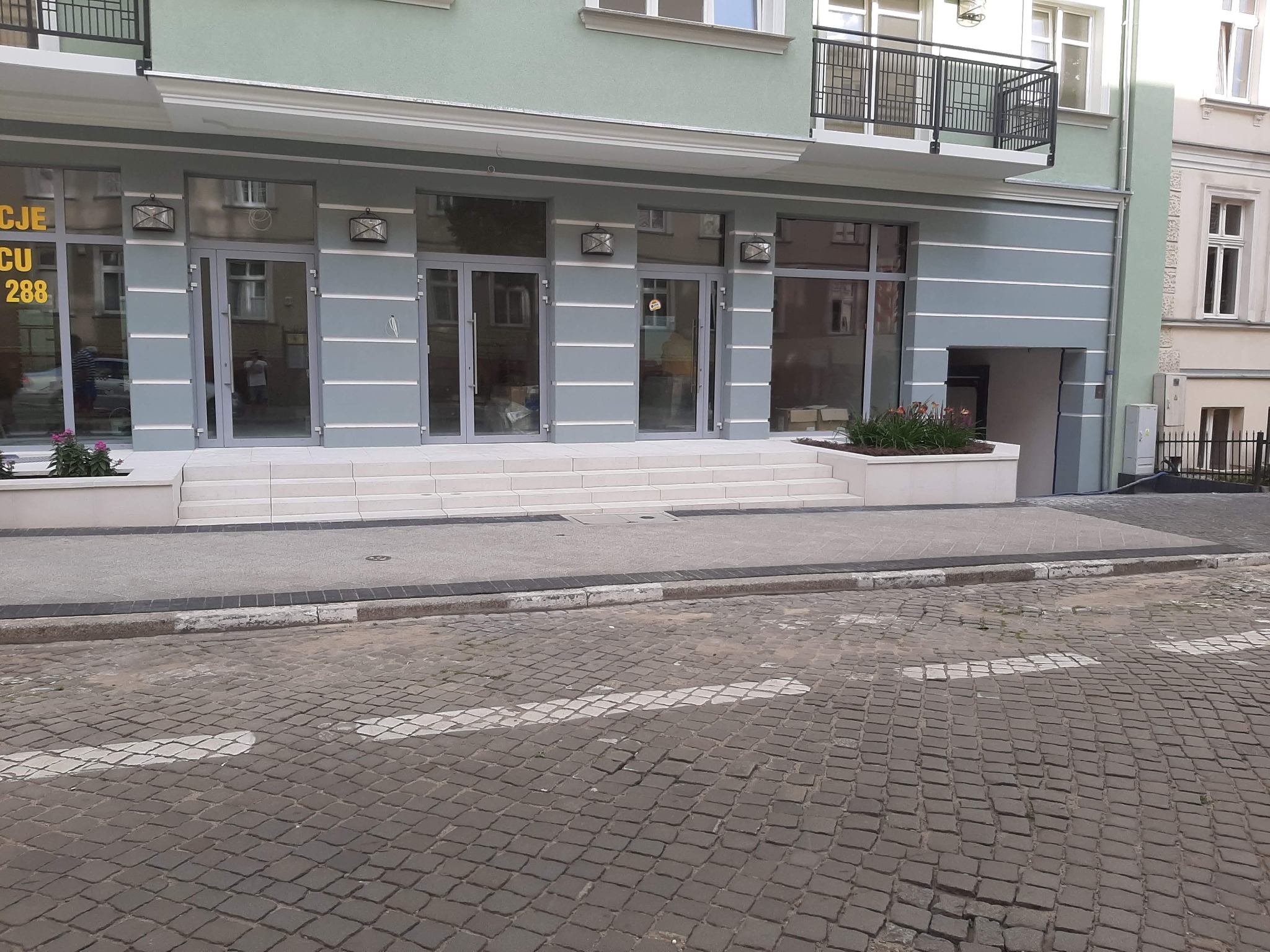 Ulica wyłożona szarą kostką brukową prowadzi do wejścia budynku z jasnoszarą elewacją, trzema szklanymi drzwiami i roślinami w donicach.
