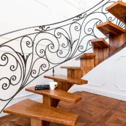 Dębowe schody wspornikowe z dekoracyjną, kutą balustradą, na jednym ze stopni leży książka i biały kubek, parkiet ułożony w geometryczny wz&oacute;r.