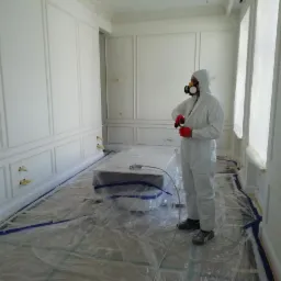 Pracownik w kombinezonie ochronnym i masce maluje natryskowo białe ściany z dekoracyjnymi panelami w jasnym pomieszczeniu, zabezpieczone folią podłogi i okna.