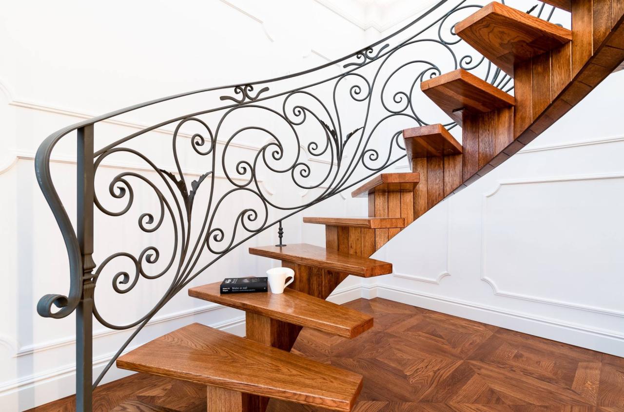 Dębowe schody wspornikowe z dekoracyjną, kutą balustradą, na jednym ze stopni leży książka i biały kubek, parkiet ułożony w geometryczny wzór.