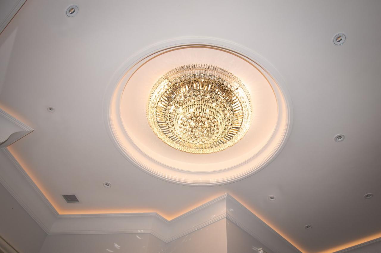 Kryształowy żyrandol w okrągłej wnęce sufitowej, otoczony profilowanym gzymsem z subtelnym podświetleniem LED, uzupełniony punktowymi lampami halogenowymi.
