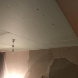 ArturCielas - Montaż sufitu podwieszanego z płyt kartonowo-gipsowych, widoczne wkręty, fragment ściany z nałożoną warstwą gładzi, prowizoryczne oświetlenie w postaci żarówki na kablu.