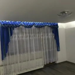 Okno z założonymi, długimi, białymi firanami z delikatnym haftem i niebieskimi zasłonami z ozdobną falbaną, widoczny grzejnik i szara podłoga.