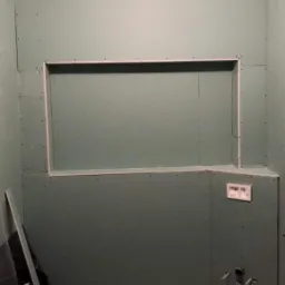 Łazienka w trakcie remontu: ściany wyłożone zieloną płytą kartonowo-gipsową z wnęką, widoczne rury instalacji wodnej, fragment wanny i poziomicy na podłodze.