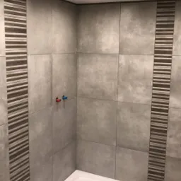 Wykończona łazienka z szarymi, kwadratowymi płytkami, dekoracyjnymi pasami z poziomych pasków i białą, narożną kabiną prysznicową.