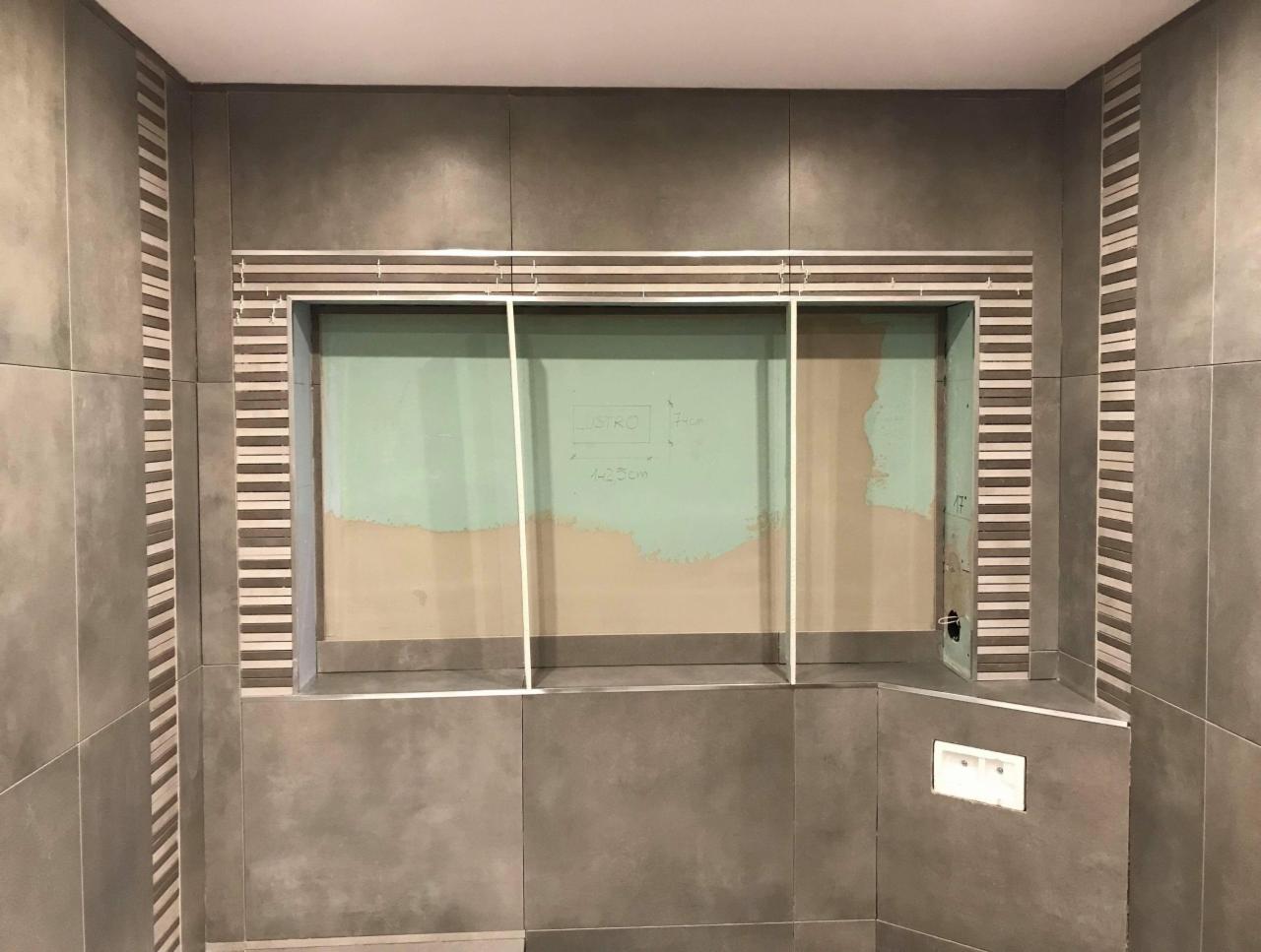 Szara łazienka w trakcie wykańczania, widoczne płytki na ścianach i obudowa wnęki z listwami dekoracyjnymi, ściana wewnątrz wnęki przygotowana pod dalsze prace.