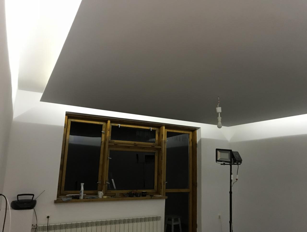 Pokój po remoncie z nową, gładką, białą powierzchnią sufitu podwieszanego, drewnianą ramą okna i lampą studyjną na statywie, widoczny grzejnik.