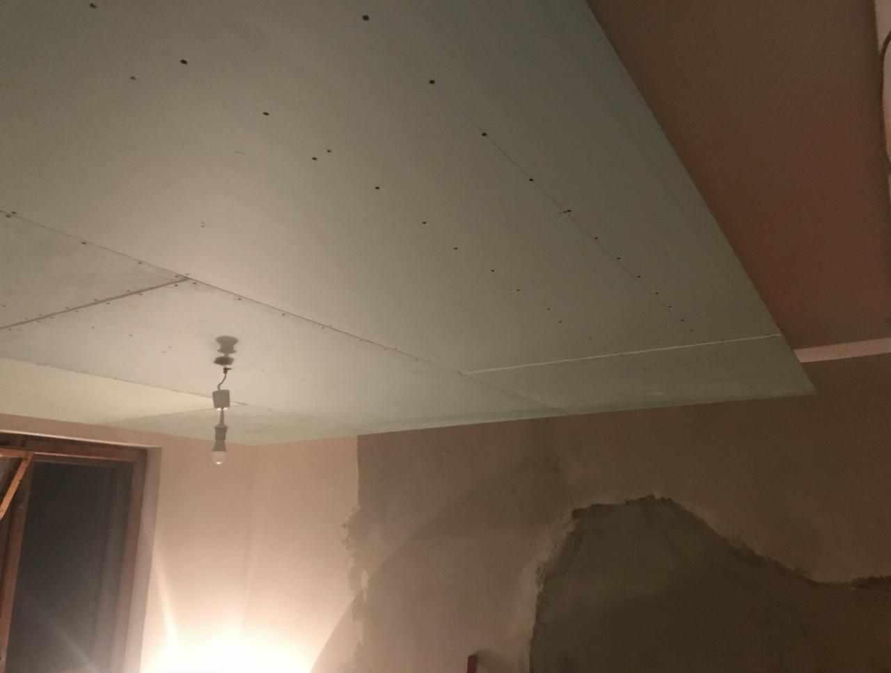 Montaż sufitu podwieszanego z płyt kartonowo-gipsowych, widoczne wkręty, fragment ściany z nałożoną warstwą gładzi, prowizoryczne oświetlenie w postaci żarówki na kablu.