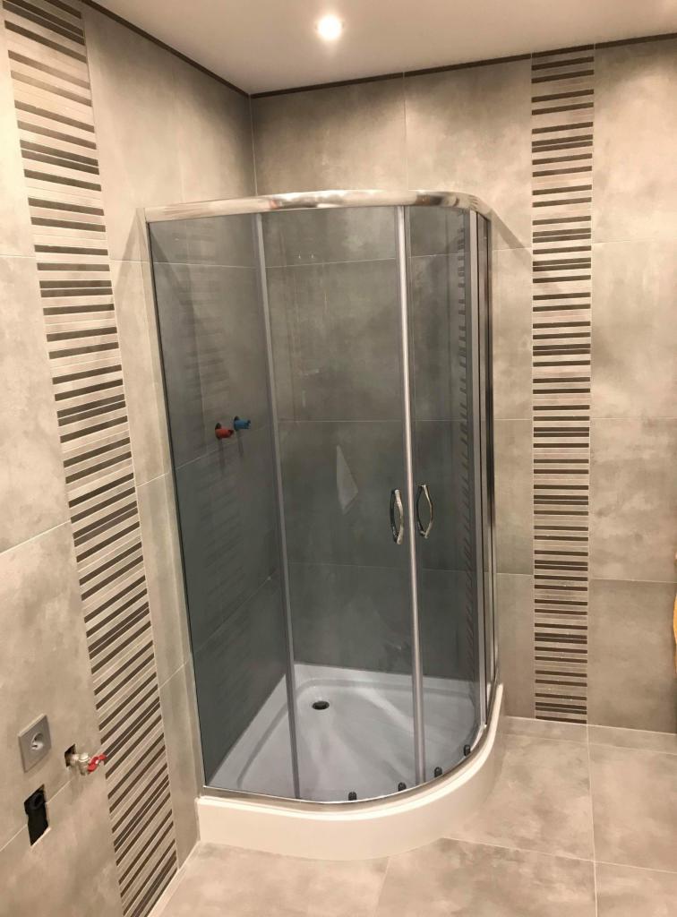 Szara łazienka z narożną kabiną prysznicową z chromowanymi profilami i szklanymi drzwiami, obok pionowy pas płytek dekoracyjnych z poziomymi paskami.