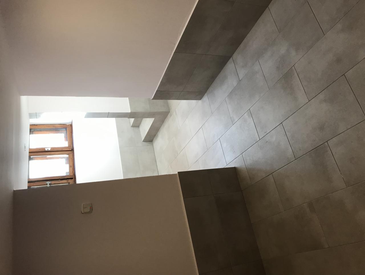 Wykończona łazienka z szarymi, prostokątnymi płytkami podłogowymi i ściennymi, widoczne fragmenty okna z drewnianą ramą i ściany w kolorze ecru.