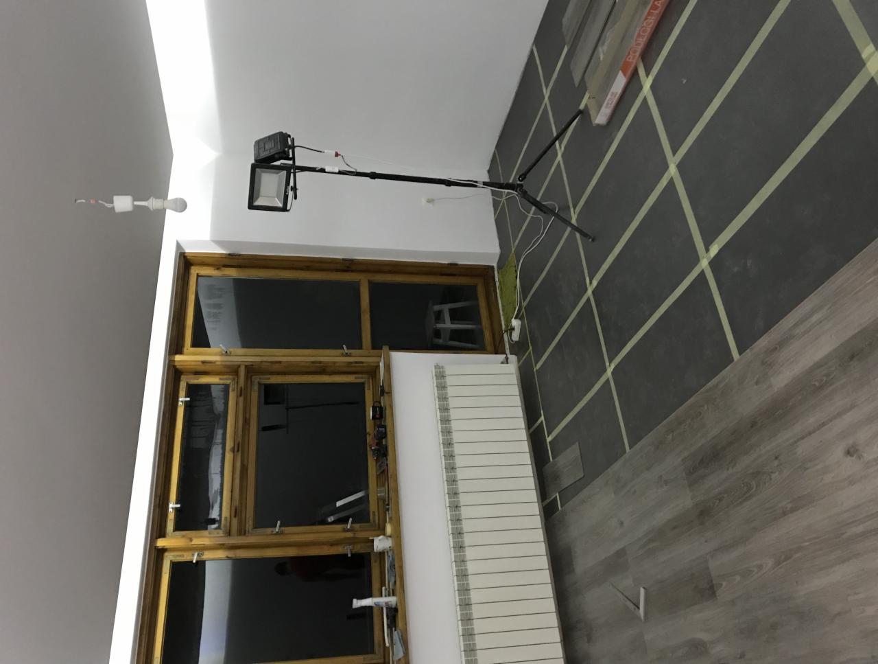 Pokój w trakcie remontu: ciemna ściana z taśmą malarską w ukośną kratę, drewniana rama okienna, grzejnik, lampa oświetleniowa na statywie i panele podłogowe.