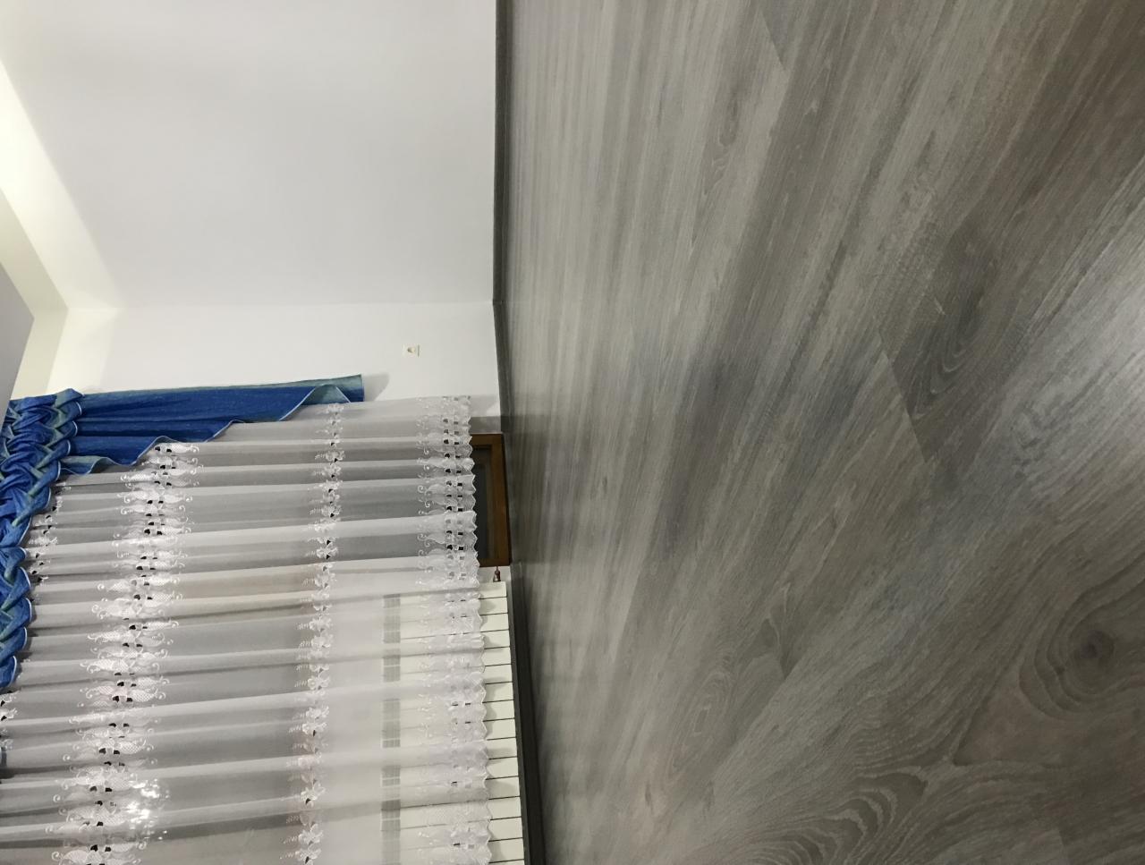 Szare panele podłogowe ułożone wzdłuż ściany, widoczne okno z białą roletą rzymską z delikatnym wzorem i niebieską dekoracją u góry.