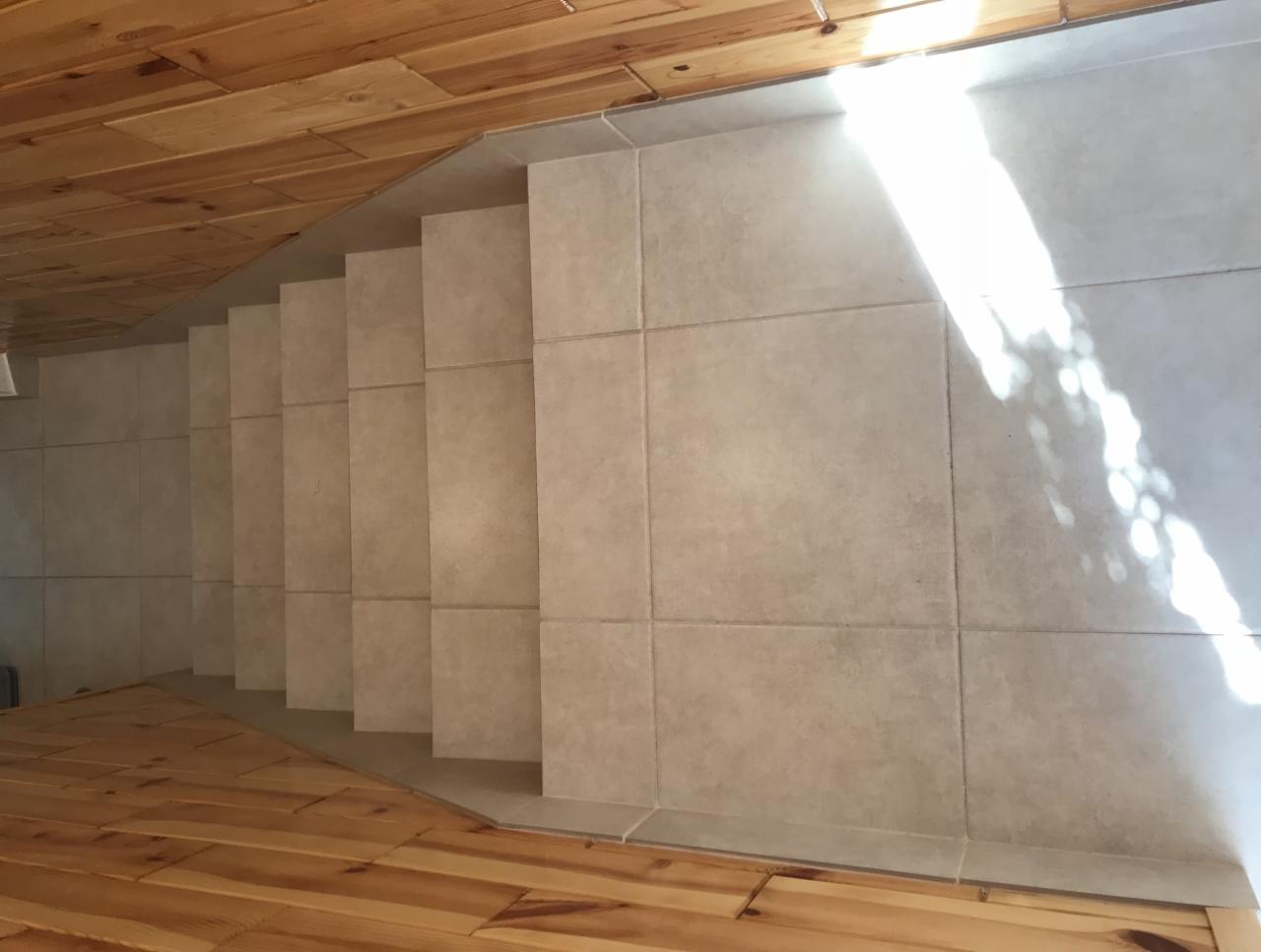 Wykończone schody z beżowych płytek ceramicznych, widok z góry, drewniana podłoga i sufit w tle, jasne światło słoneczne.